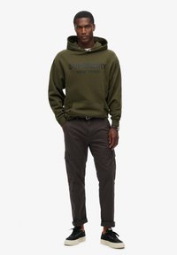 Sweat-shirt à capuche vert olive avec logo noir, poche frontale, associé à un pantalon cargo sombre et des baskets noires, avec des manches retroussées.
