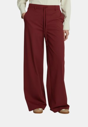 Persona che indossa pantaloni bordeaux a gamba larga con tasche frontali e cintura annodata in vita, abbinati a scarpe beige e top a maniche lunghe color crema.
