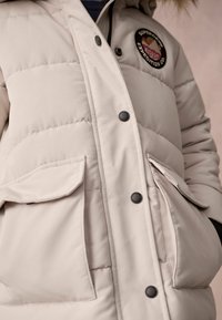 Beige Pufferjacke mit einem gesteppten Design, ausgestattet mit zwei Klappentaschen, schwarzen Druckknöpfen und einem runden Patch-Logo auf der Brust.