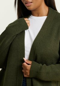 Kvinna som bär en mörkgrön stickad kofta över en vit skjorta, håller koftans kanter med båda händerna nära bröstet.