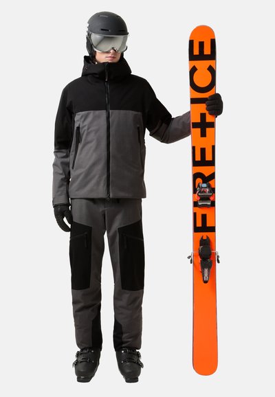 Lyžařský outfit s černou a šedou bundou, šedými kalhotami a černými rukavicemi. Drží oranžové lyže s černým nápisem a vázáním. Černá helma a brýle.