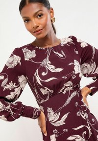 Vrouw met glad, donker haar in een staart, die gouden knoop oorbellen en een kastanjebruine jurk met witte bloemenpatronen draagt, handen op de heupen.