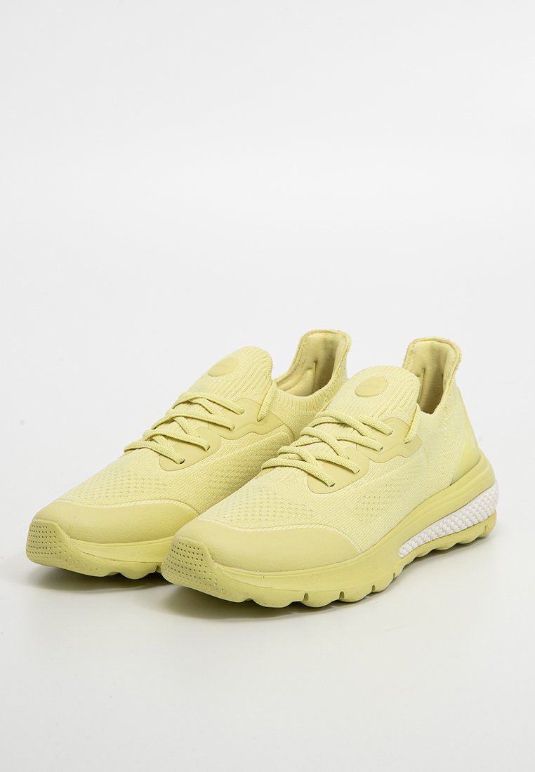 Un par de zapatillas de correr de punto color amarillo claro con cordones en la parte delantera y suelas blancas acolchadas sobre un fondo blanco.