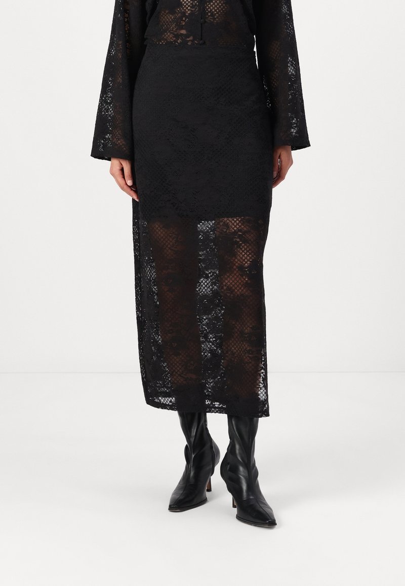 Femme portant une jupe midi en dentelle noire avec une doublure opaque et des bottes à talons en cuir noir à bout pointu, debout devant un fond blanc.