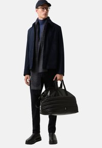 Mężczyzna w granatowej kurtce, szaliku i czapce trzymający czarną, pikowaną torbę sportową, stojący na tle jednolitego białego tła.