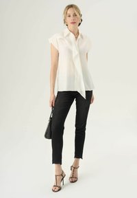Blusa bianca a maniche corte con dettaglio arricciato sul davanti, abbinata a jeans neri e sandali con cinturini leopardati; accessorata con una piccola borsa nera.