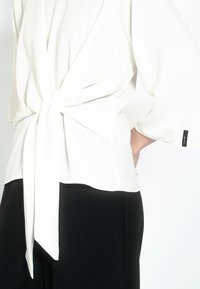 Blusa blanca ligera con un lazo en la parte trasera y mangas tres cuartos. Presenta una textura suave y un discreto acento de logo negro.