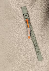 Gros plan sur un tissu polaire beige avec une fermeture éclair verte et une languette orange portant l'inscription « SUBLEVEL ».