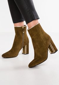 Bottines en suede marron avec un bout carré, une fermeture éclair sur le côté et un accent géométrique en or sur le talon bloc.