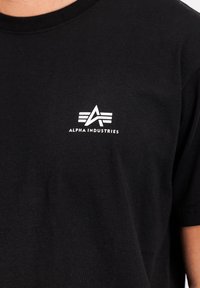 T-shirt en coton noir avec un logo "Alpha Industries" blanc et un design stylisé de "A" sur la partie gauche de la poitrine. Manches courtes, encolure ronde.