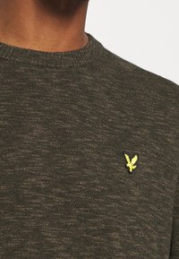 Tummanvihreä neulepaita pyöreällä pääntiellä; rinnan kohdalla pieni keltainen kotkanlogo ja hienovarainen tekstuuri kauttaaltaan.