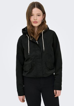 Jeune femme aux longs cheveux bruns, portant des boucles d'oreilles anneaux, une veste noire à capuche avec des cordons blancs, et un pantalon noir sur un fond uni.