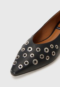 Zapato de cuero negro con punta afilada, adornado con embellishments de ojales plateados. El interior presenta un forro suave en tono marrón claro.