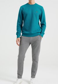 Tyrkysová mikina s žebrovanou strukturou a kulatým výstřihem, v kombinaci s kalhotami v kostkovaném vzoru v černobílé barvě a bílými teniskami.