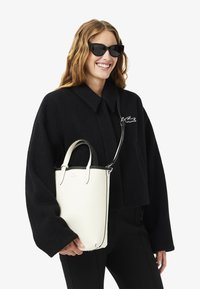 Sac à main en cuir texturé blanc avec des accents noirs, poignées arrondies et une bandoulière amovible. Le modèle porte une grande veste noire.