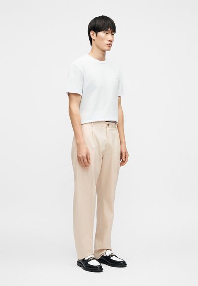 Λευκό βαμβακερό t-shirt με κοντά μανίκια, συνδυασμένο με ανοιχτό μπεζ παντελόνι. Το υπόδημα είναι από μαύρο δέρμα με λευκή λεπτομέρεια και αλυσιδάκι.