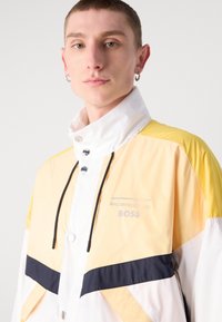 Geacă de vânt galbenă și albă cu accente negre, având un guler înalt, butoane cu capse și logo-ul "Porsche BOSS". Material neted, ușor.