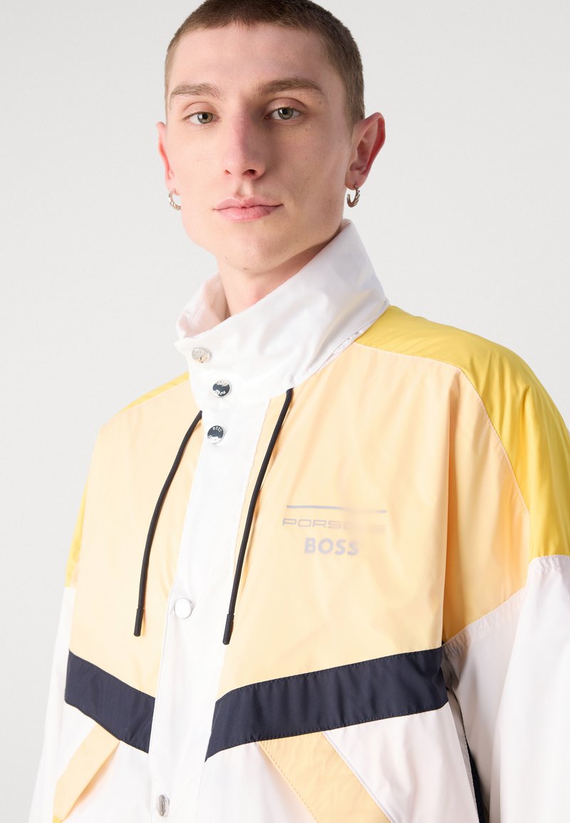 Veste coupe-vent jaune et blanche avec des accents noirs, dotée d'un col haut, de boutons-pression et du logo "Porsche BOSS". Tissu lisse et léger.