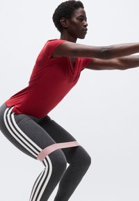 Άτομο με κόκκινο πουκάμισο και γκρι κολάν με λευκές ρίγες κάνει κάθισμα με λάστιχο αντίστασης πάνω από τα γόνατα, μπροστά σε απλό φόντο.