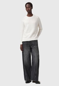 Donna che indossa un maglione bianco a trecce, jeans neri svasati sbiaditi e ballerine nere con fibbie dorate, in piedi contro uno sfondo neutro.