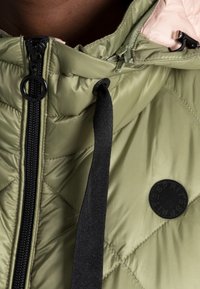 Zielona kurtka puchowa z diamentowym pikowaniem, czarnym zamkiem błyskawicznym i naszywką z logo. Wnętrze jest różowe, a kurtka ma regulowany czarny ściągacz.