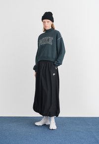 Tumši zaļš Nike džemperis ar baltu logo, apvienots ar melnu, plūstošu svārku un baltām sporta kurpēm. Modelis valkā melnu bīni.