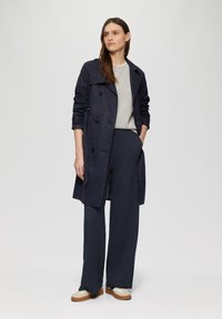 Marineblauwe trenchcoat met dubbelzijdige knopen, grijze trui en brede marineblauwe broek. Schoeisel bestaat uit bruine en witte sneakers.