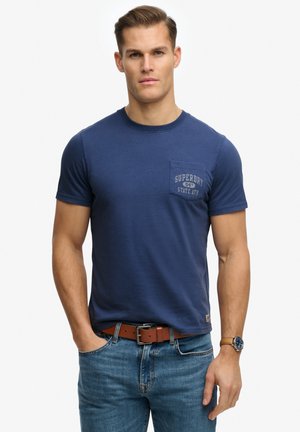 Mężczyzna w granatowym t-shircie Superdry z kieszenią, niebieskich dżinsach, brązowym skórzanym pasku i zegarku na rękę, stojący z jedną ręką w kieszeni.