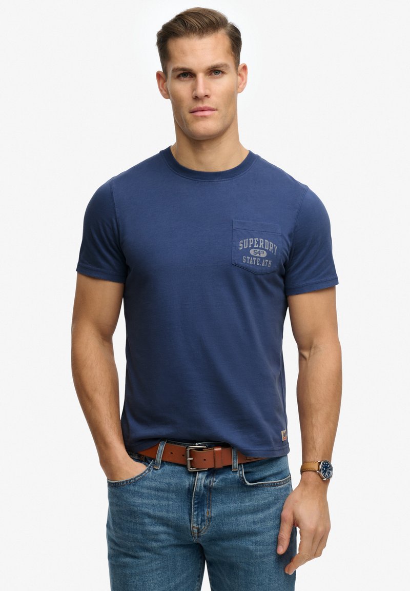 Homme portant un t-shirt Superdry bleu marine avec une poche, un jean bleu, une ceinture en cuir marron et une montre-bracelet, debout avec une main dans la poche.