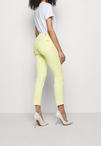 Vaqueros skinny amarillos hasta el tobillo con un dobladillo deshilachado, combinados con tacones altos beige y una camiseta blanca. Presenta bolsillos traseros sutiles.