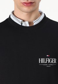Juodas megztinis su apykakle, su balta "HILFIGER" logotipu ir mažu raudonai-mėlynu-baltu emblema, uždėtas ant šviesiai mėlynos raštuotos marškinių.