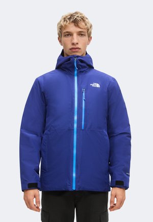 Młoda osoba w niebieskiej kurtce The North Face z kapturem, zamkiem błyskawicznym z przodu i suwakiem na kieszeni piersiowej, stoi na jednolitym tle.