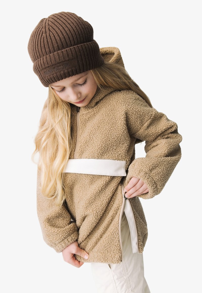 Brun fleece hoodie med hætte og hvid rem. Modellen trækker i en lynlås på siden og bærer en strikket brun hue. Langt, lige hår er synligt.