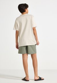 Ljusbeige polo skjorta med korta ärmar, ihop med texturerade gröna shorts. Bärs med svarta sandaler. Bakifrån mot en neutral bakgrund.