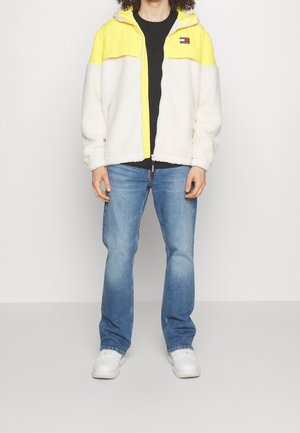 Osoba ubrana w żółto-białą bluzę polarową na zamek, czarną koszulkę, niebieskie dżinsy i białe trampki, stojąca na jednolitym białym tle.