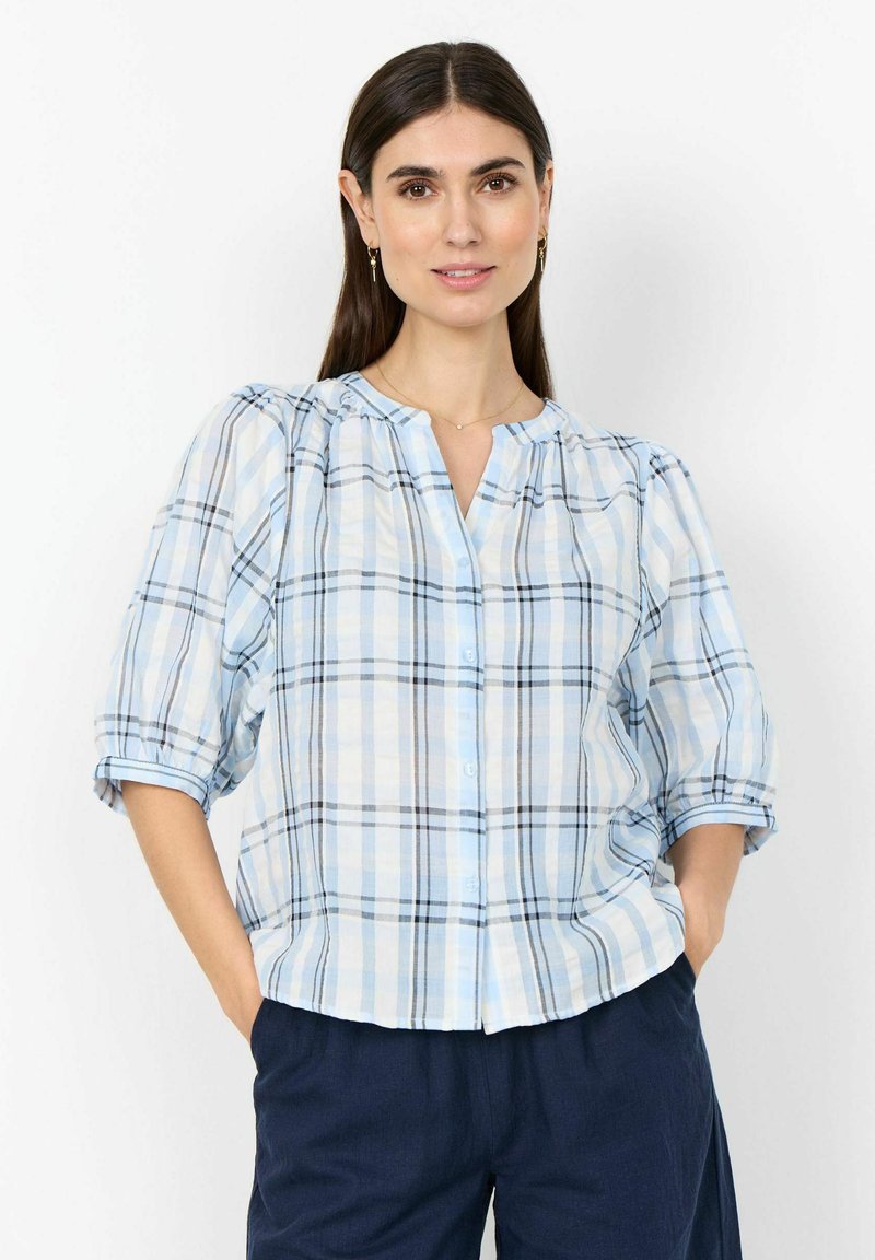 Femme portant une chemise à carreaux bleu clair et blanc avec des manches bouffantes et un pantalon bleu marine, debout les mains dans les poches.