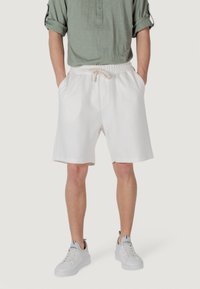 Un uomo sta in piedi indossando una camicia a maniche lunghe verde chiaro e pantaloni corti bianchi, abbinati a scarpe da ginnastica bianche, su uno sfondo neutro.