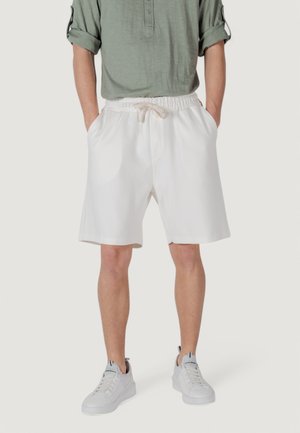 Un uomo sta in piedi indossando una camicia a maniche lunghe verde chiaro e pantaloni corti bianchi, abbinati a scarpe da ginnastica bianche, su uno sfondo neutro.