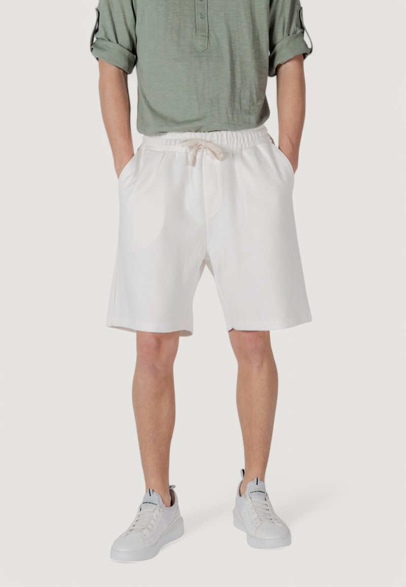 Un uomo sta in piedi indossando una camicia a maniche lunghe verde chiaro e pantaloni corti bianchi, abbinati a scarpe da ginnastica bianche, su uno sfondo neutro.