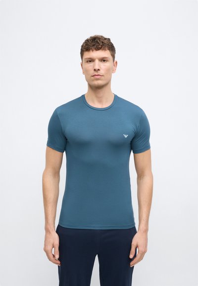 Άνδρας που στέκεται μπροστά φορώντας εφαρμοστό μπλε-πράσινο κοντομάνικο πουκάμισο και σκούρο παντελόνι, μπροστά σε απλό λευκό φόντο.