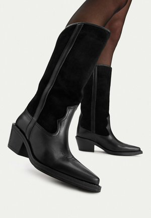 Stivali neri in pelle e camoscio al ginocchio con punte appuntite e tacchi a blocco indossati su gambe contro uno sfondo semplice.