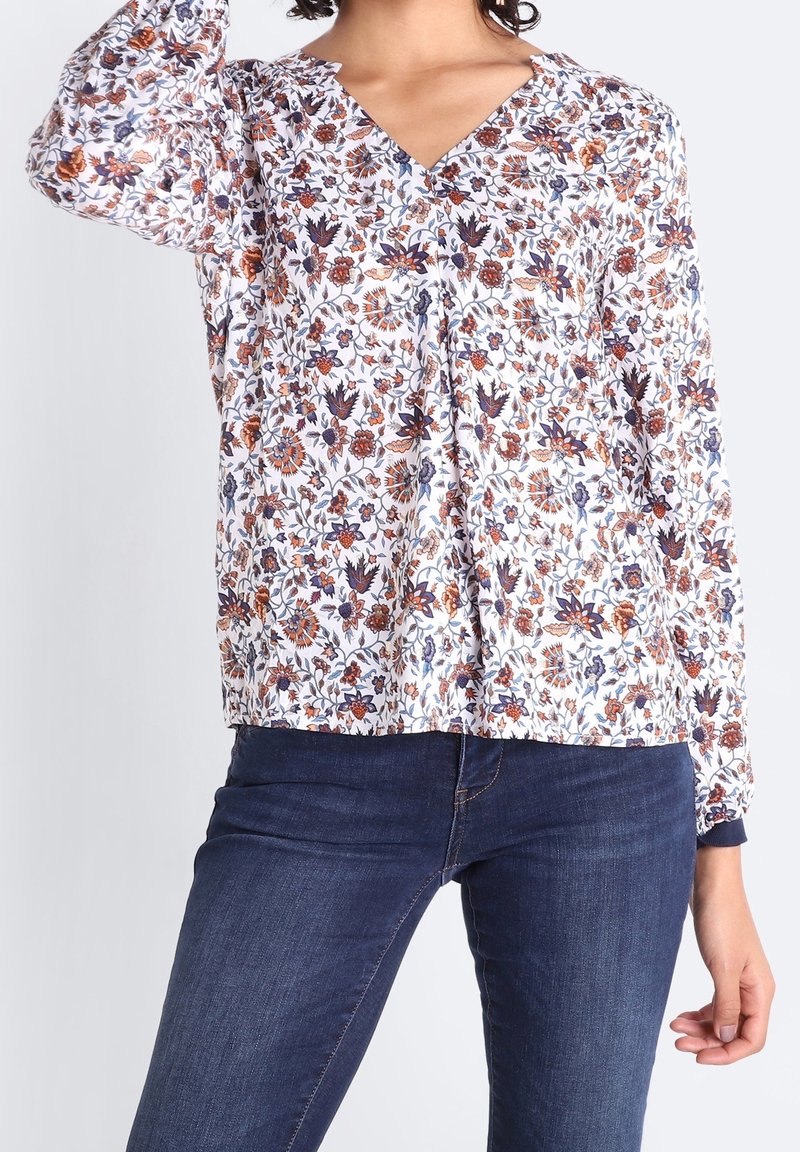Femme portant un chemisier blanc à fleurs bleues, orange et marron avec manches longues, associé à un jean bleu foncé.