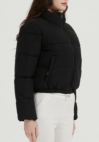 Veste courte en duvet noire avec col montant, fermeture éclair et deux poches latérales. Caractérisée par des sections matelassées et un ourlet et des poignets élastiques.