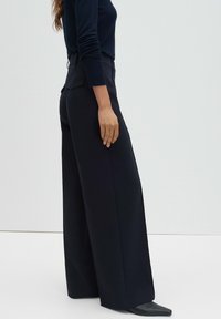 Pantalon noir à jambes larges avec une taille haute, tissu lisse et deux poches arrière. Associé à un haut sombre ajusté et des chaussures noires pointues.