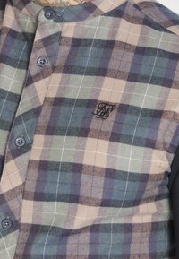 Camicia a quadri con bottoni in beige, verde e blu navy. Presenta un colletto alla mandarina, maniche nere e un logo ricamato sul petto.