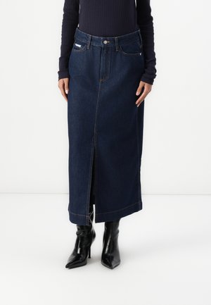 Femme portant une jupe midi en denim bleu foncé avec une fente devant, des bottines noires à talons pointues, et un top à manches longues côtelé bleu marine.