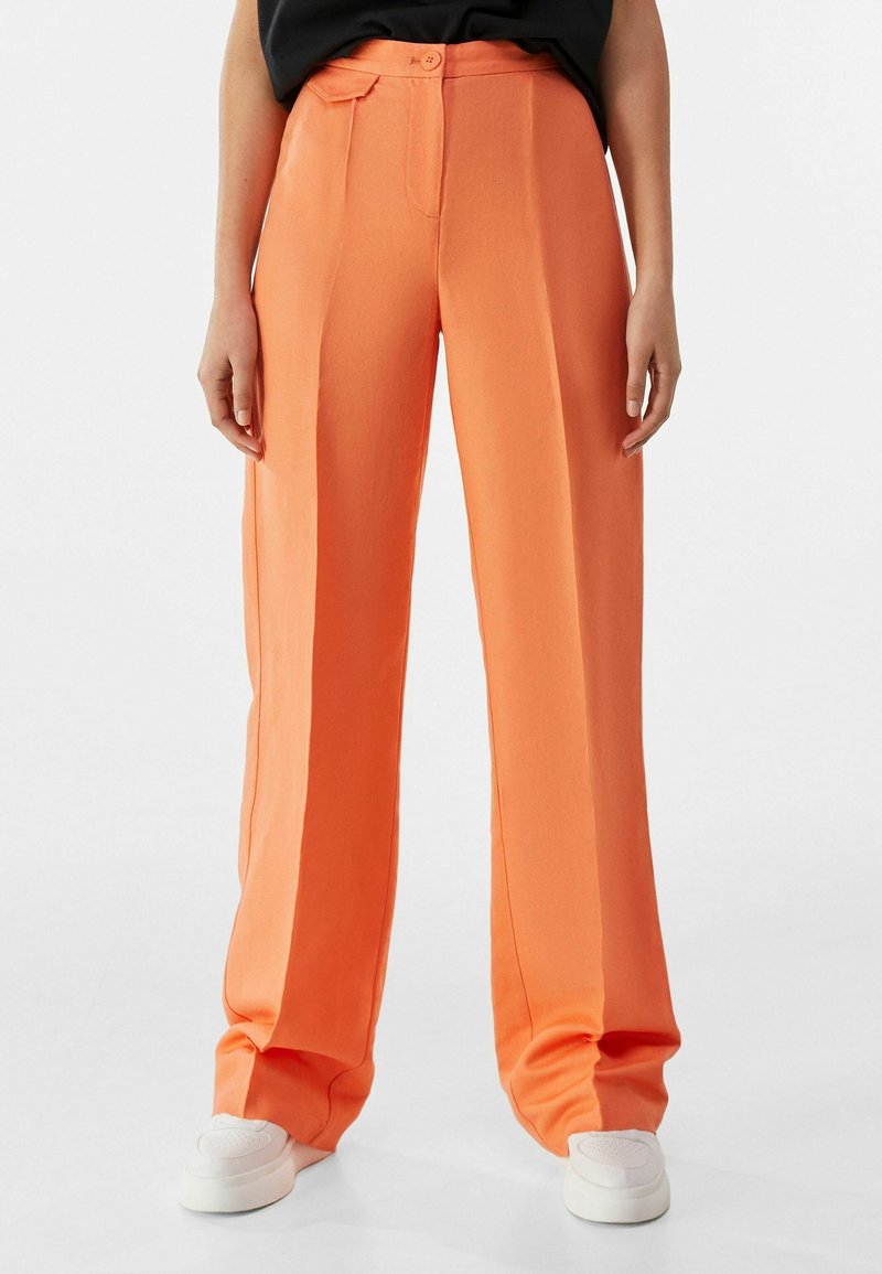 Pantalones de pierna ancha en color naranja, fabricados en tejido suave, con cierre de botón y pliegues laterales, combinados con zapatillas blancas.