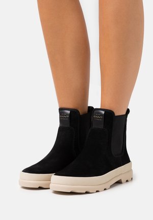 Bottines en daim noir avec semelles en caoutchouc beige portées sur des jambes nues, équipées de panneaux latéraux élastiques et d'une languette de traction avec logo.