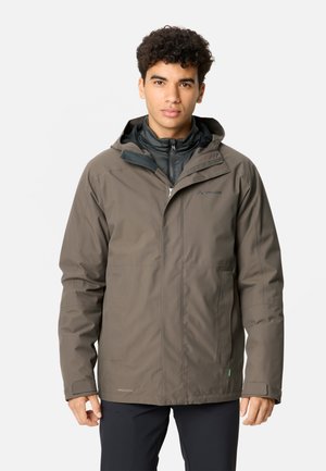 Man draagt een bruine waterdichte capuchonjas over een zwart vest met rits, kijkt recht vooruit met een neutrale gezichtsuitdrukking.