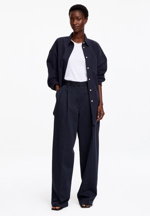 Mujer con pantalones anchos azul marino, camisa grande a juego abotonada, camiseta blanca y tacones abiertos negros, de pie con las manos en los bolsillos.